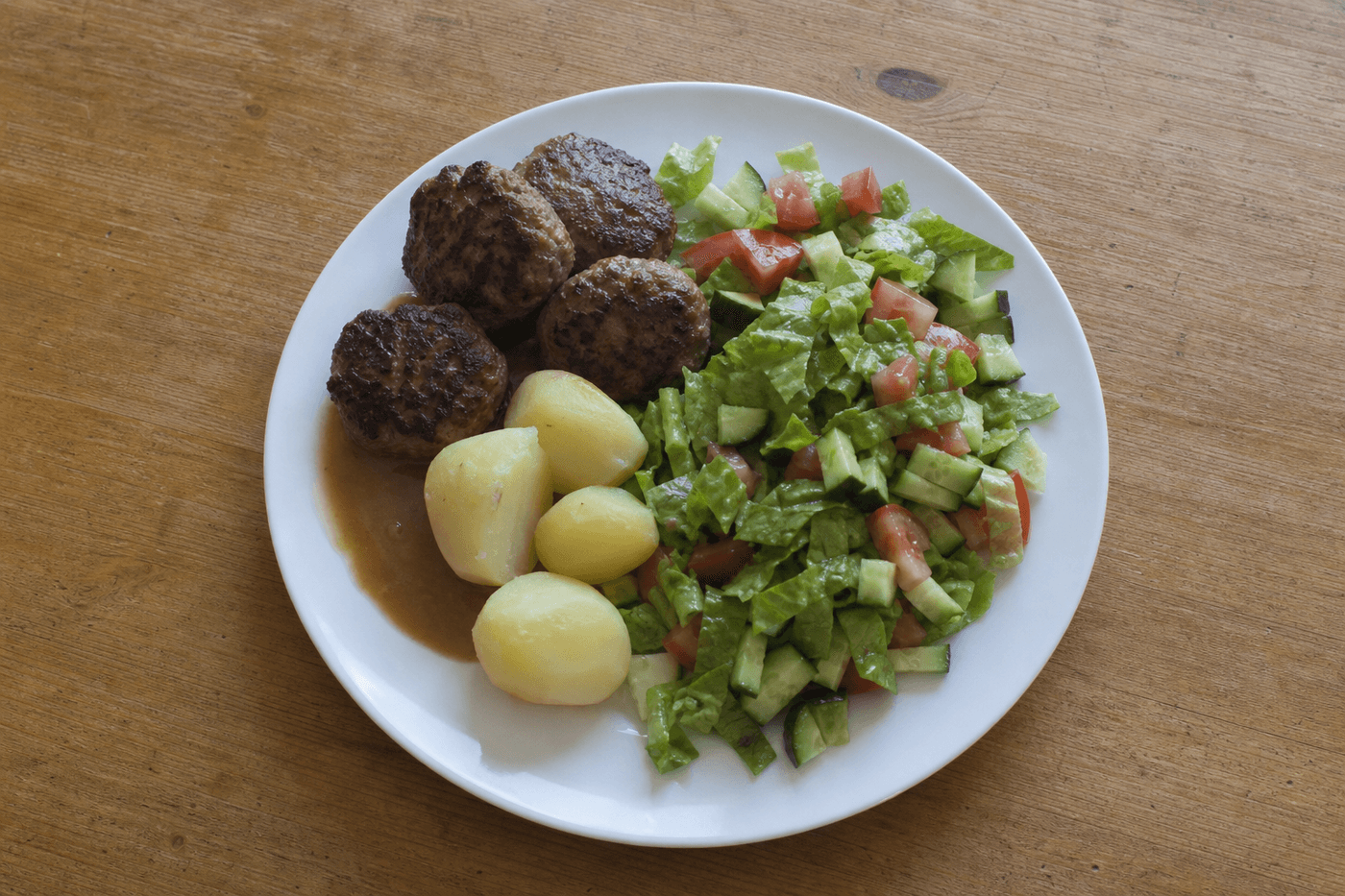 Frikadeller, kartofler, brun sovs og en stor salat på en hvid tallerken på et køkkenbord i eg